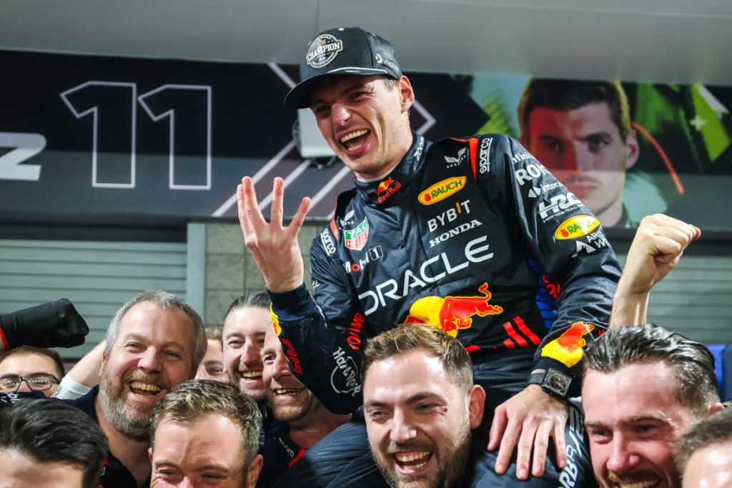 Max Verstappen celebrating with the Red Bull Racing team, held aloft and flashing four fingers after winning the Formula 1 championship.